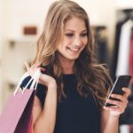 Making the retail connection. Beautiful young woman with shopping bags using her smart phone with smile while standing at the store